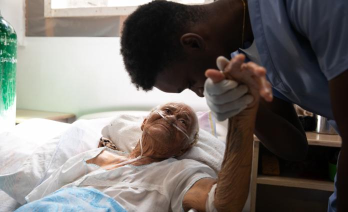 An elderly Haitian woman lays in a hospital bed in the internal medicine department. A young male Haitian doctor holds her hand and leans over to talk to her.