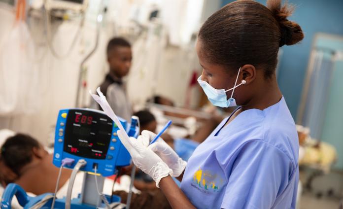 A female clinician wearing light blue scrubs, blue medical mask, and white exam gloves writes on a piece of paper in an emergency room. She stands in front of a vital signs monitor. In the background, patients lie on hospital beds.