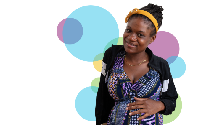 A pregnant Haitian woman smiles at the camera with her head tilted slightly to the left. Her right hand is on her stomach. Her dark hair is braided and she is wearing a yellow headband, a black cardigan, and a purple, black, and white printed dress.