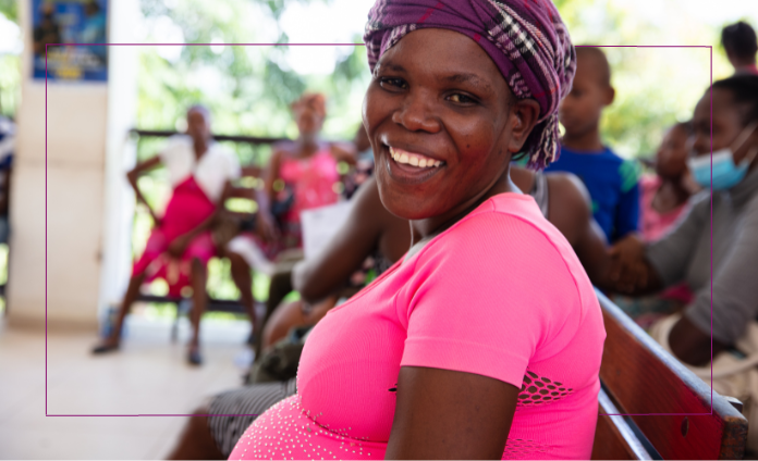 A pregnant Haitian woman wearing a neon pink top and a purple plaid head wrap turns her head and smiles brightly at the camera.