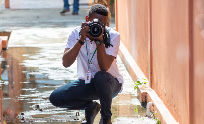 A young Haitian man crouches outsided. He looks through the viewfinder of a large camera, which is pointed at the viewer. The paved ground is wet from rain.