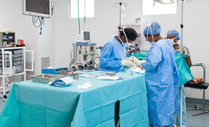 A team of Haitian doctors perform a surgical procedure. They bend over a large surgical table draped in a teal cloth. The surgeons wear light blue scrubs, medical masks, and caps.