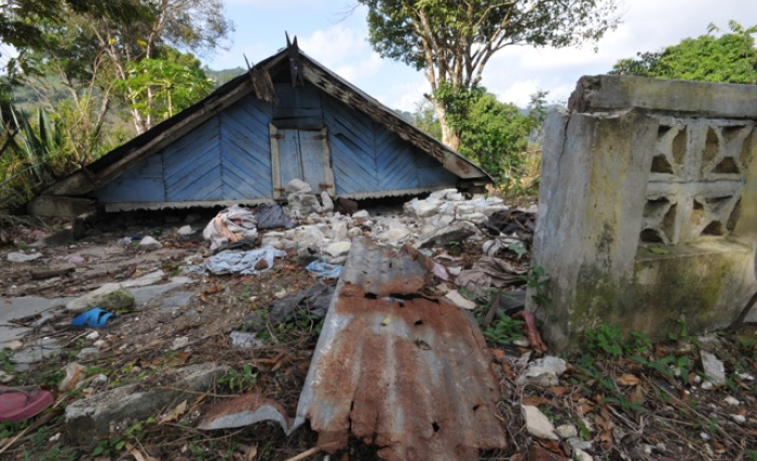 In the mountains, the wooden and tin roof of a house sits on the ground behind rubble.