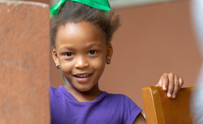 A young girl smiles cheerfully at the camera. She wears a purple T-shirt and a green bow in her hair.