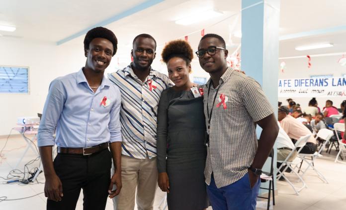 Four Haitians in business casual clothes stand together in a large, bright room, smiling and looking relaxed.