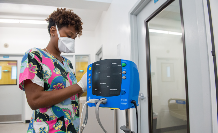 In a hospital ward, a Haitian woman in bright patterned scrubs, N-95 mask, & gloves adjusts a monitoring machine.