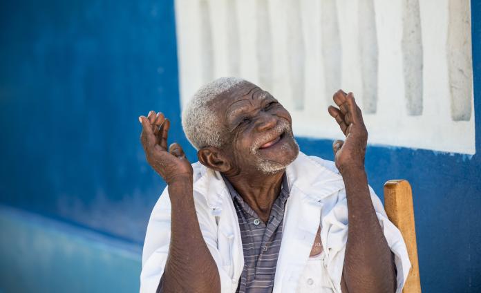 Elderly Haitian man smiling and gesturing with his arms in front of a blue wall at Kay Gran Moun.