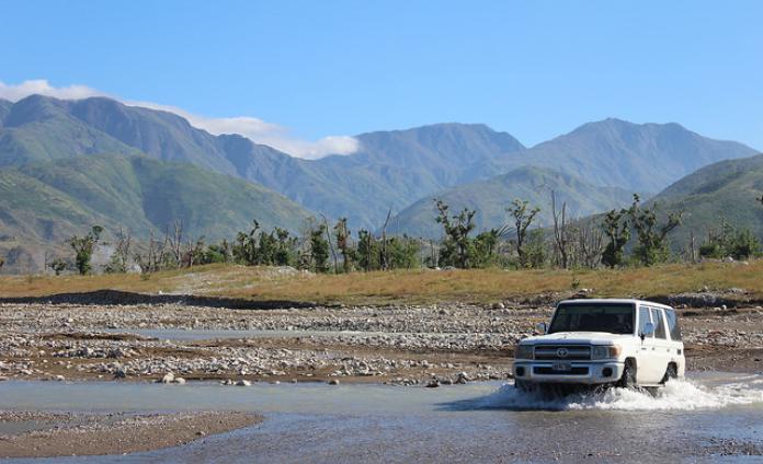 A white ambulance drives through a shallow river. There is a grassy plain and mountains in the background.