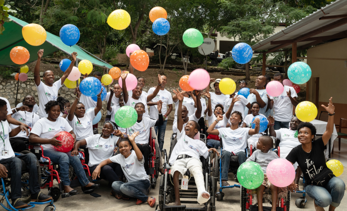 Group photo of over 2 dozen Haitians laughing and tossing colorful balloons in the air. Many are in wheelchairs.