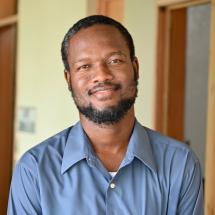 Headshot of a Haitian man smiling kindly at the camera. He is wearing a blue button-down shirt and has close-cropped hair & a short beard and mustache.