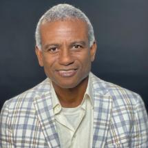 Headshot of a smiling Haitian man with short-cropped gray hair, wearing a light-colored plaid jacket over a white shirt.
