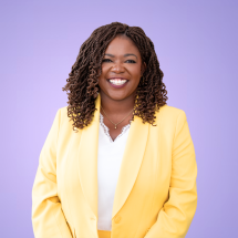 A portrait of Ruthzee Louijeune, a Haitian politician who has coiled, shoulder-length brown hair and is wearing a yellow blazer and white shirt. She smiles brightly.