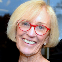 A headshot of Beth Floor smiling and wearing a black blouse and bright red glasses. 