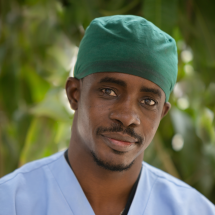 A dark-skinned man smiles at the camera. He is wearing light blue v-neck scrubs, a silver necklace, and a green surgical cap. He has a short mustache..