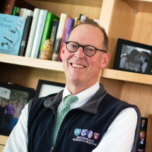 A smiling Dr. Paul Farmer stands in an office wearing glasses, a white collard shirt, a green tie, and a Brigham and Women's Hospital vest.