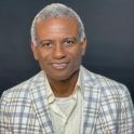 Headshot of a smiling Haitian man with short-cropped gray hair, wearing a light-colored plaid jacket over a white shirt.