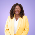 A portrait of Ruthzee Louijeune, a Haitian politician who has coiled, shoulder-length brown hair and is wearing a yellow blazer and white shirt. She smiles brightly.