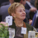 Diane Kaneb is looking to her left and wears a grey blouse, a black blazer, a scarf on one shoulder, and a gold necklace.