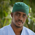 A dark-skinned man smiles at the camera. He is wearing light blue v-neck scrubs, a silver necklace, and a green surgical cap. He has a short mustache..