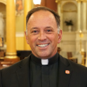 Rev. John Unni smiles and wears a black shirt and blazer with a white clerical collar.
