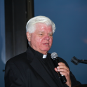 Rev. Gerald Osterman holds a microphone and wears a black shirt and blazer with a white clerical collar.