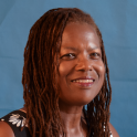 A headshot of Dr. Michele David smiling. She has locs and wears a floral blouse and is situated against a blue background.