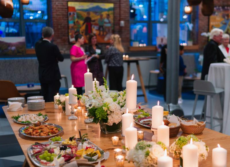 A wooden table with white candles, flower arrangements, and appetizers. In the background are people chatting and large windows. Outside, dusk is falling.