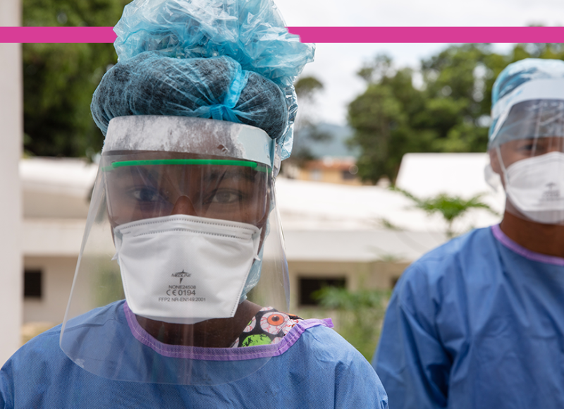 A Haitian woman clinician wears personal protective equipment during the COVID-19 pandemic. She wears a blue medical gown, a white face mask, a clear face shield, and a protective net over her hair.