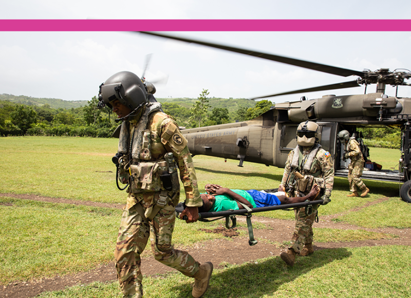 An earthquake patient is taken out of a military helicopter on a stretcher. Two military pilots in full gear carry the front and back of the stretcher across a field.