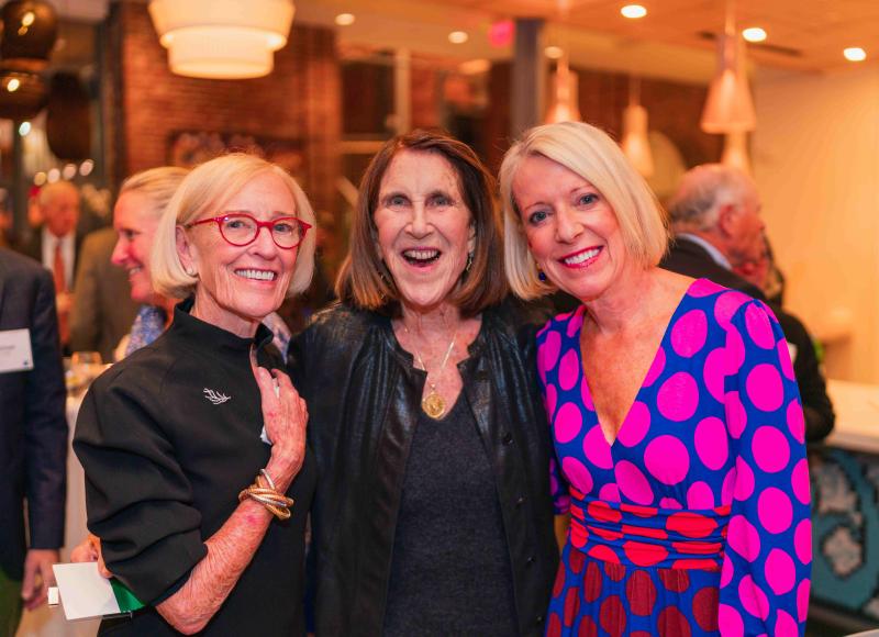 Three white women pose together with their arms around each other’s backs. They smile joyfully at the camera.