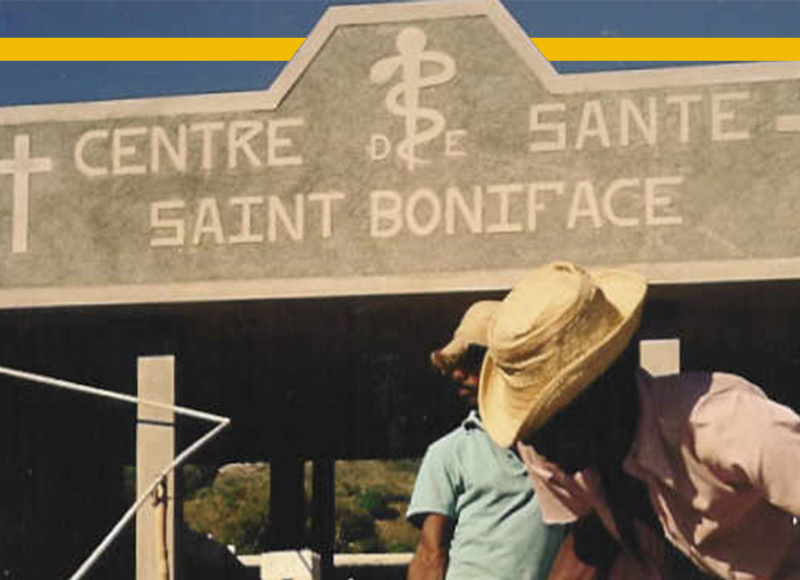 A photo of the old Saint Boniface health center with two Haitian men digging a hole in front of the entrance.