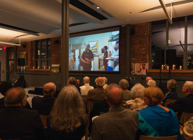 In a large, darkened room a seated crowd of adults watches a video on a large screen. 