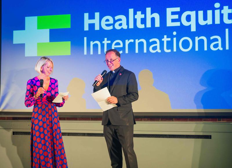 A white woman in a brightly-colored dress and a white priest in a black suit stand next to each other, holding microphones, in front of a large screen with the HEI logo.