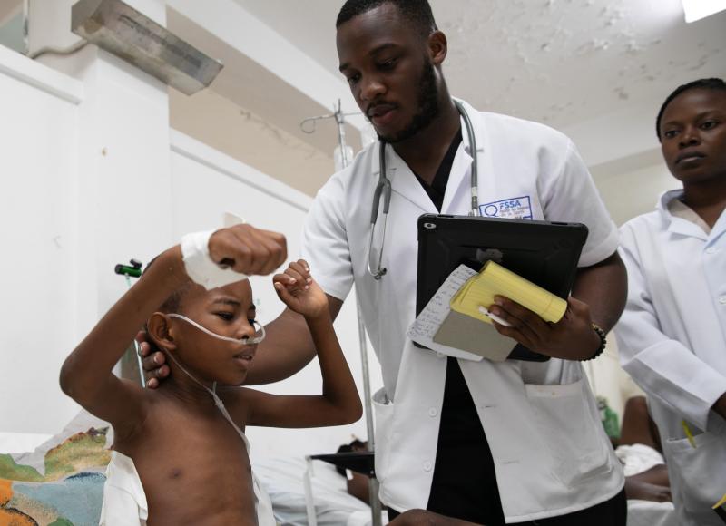 A young Haitian boy sitting on a hospital bed, raising his arms. He has tubes on his face for oxygen and a bandage and tube on his right side. A Haitian man in a white lab coat touches the boy’s neck.