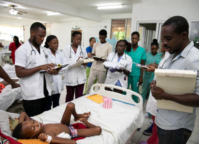 Ten Haitian men and women, most in white lab coats and with stethoscopes around their necks, stand around a young boy’s hospital bed, holding clipboards. The boy wears blue trunks and has a bandage on his right side.