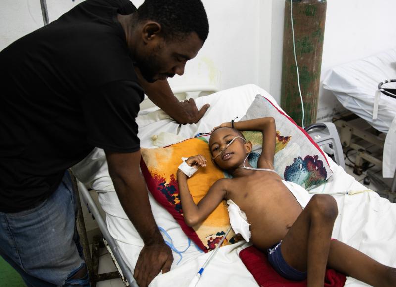 A Haitian man in a black T-shirt leans protectively over a hospital bed, looking at the young boy resting there with a bandaged side.