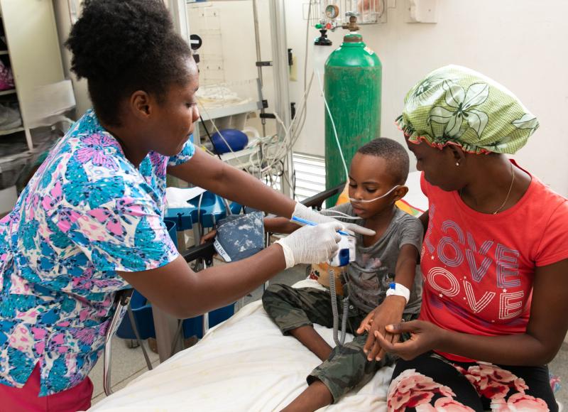 A Haitian woman in scrubs with a pink and aqua butterfly design holds a medical device in front of a young boy’s chest. The boy wears a gray T-shirt and camo pants. His mother, in a red T-shirt, looks down at him lovingly and holds his hand.