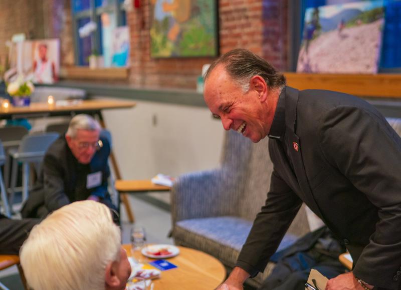 Two men shake hands over a small table. The man on the left is standing and is wearing a priet's collar. The man on the left is seated and seen from behind.