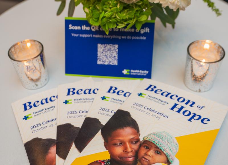 A small event table decorated with a short vase of flowers, two votive candles, a blue postcard, and four event programs