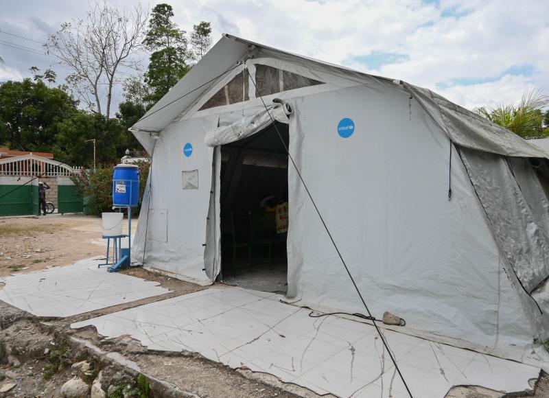 Exterior view of a large light grey tent set up in a courtyard. At one corner is a handwashing station with water tank and bucket.