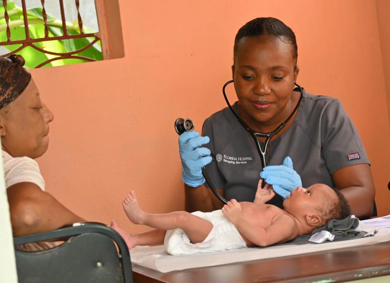 A Haitian woman doctor with a stethoscope smiles at an active baby on the table in front of her. A light-skinned Haitian woman sits close by.