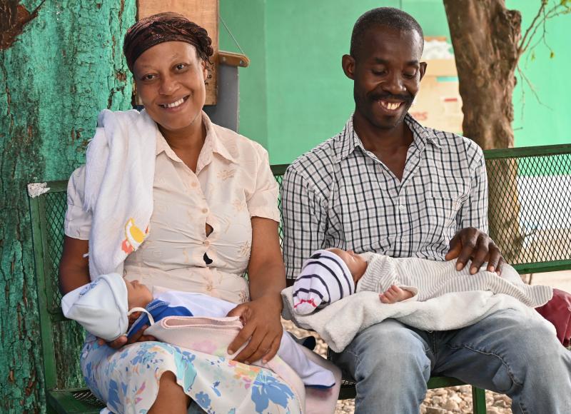 Relaxed and smiling, a light-skinned Haitian woman and dark-skinned Haitian man sit outside on a green metal bench, holding their babies on their laps.
