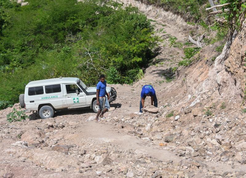 A white 4x4 vehicle marked “ambulance” is parked at the bottom of a steep, rocky hill while 2 men in blue T-shirts shift boulders.