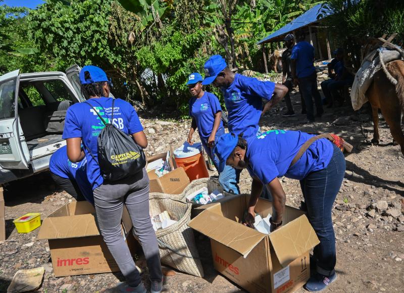 On a dirt/gravel road, 6 Haitians in bright blue HEI T-shirts and caps unload cardboard boxes of supplies from a white 4x4.