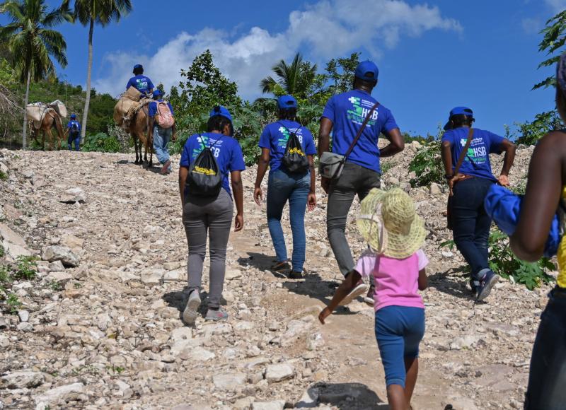 A group of Haitians in blue HEI T-shirts and caps, along with a small girl in pink and two loaded mules, reach the top of a rocky hill under blue sky.