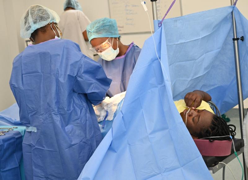 A Haitian woman in braids lies on an operating table, flanked by Haitian surgeons in full PPE.