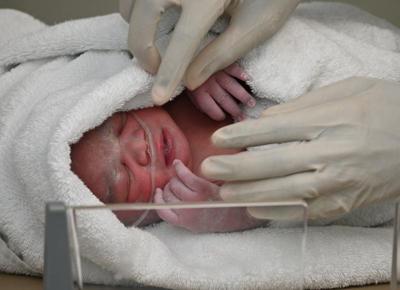 Close-up photo of a newborn infant with an oxygen tube, swaddled in a white towel. The newborn’s tiny pink hands contrast with the gloved hands of a nurse.