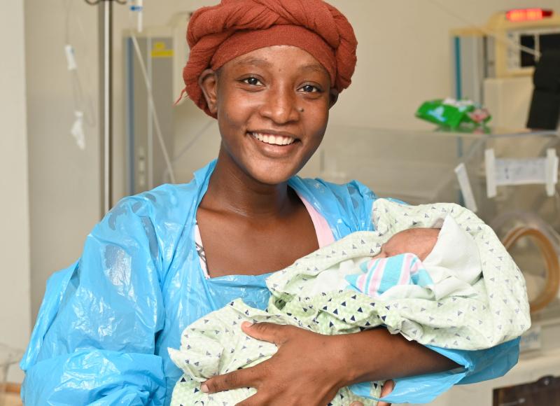 A young Haitian woman wearing a orange-brown head wrap and blue hospital gown smiles happily. She holds a small infant in her arms.