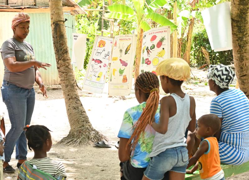 Outside, in front of sunny vegetation, a Haitian woman in a head wrap, T-shirt, and jeans speaks to an attentive group of women and children. Educational posters hang nearby.