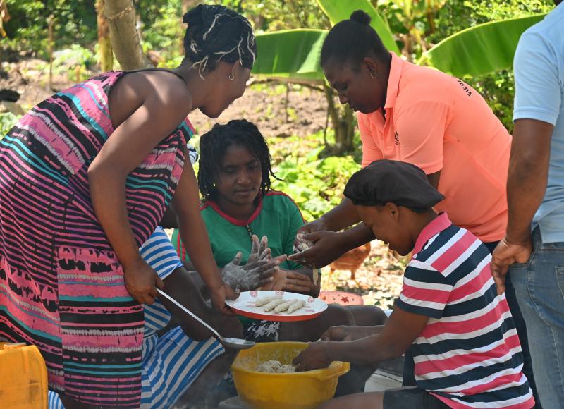 In a shady outside area, 5 Haitian women form a loose circle around a bowl of dough they’re forming into long dumplings. Some wear colorful polo shirts and others striped dresses.